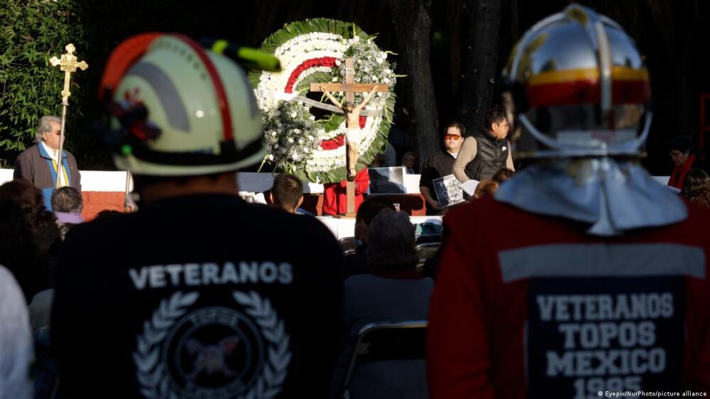 Dos bomberos vetaranos, con una referencia expresa al terremoto de 1985 en sus uniformes, atienden la ceremonia religiosa, con un cristo al fondo y una corona de flores en recuerdo a las vítimas.