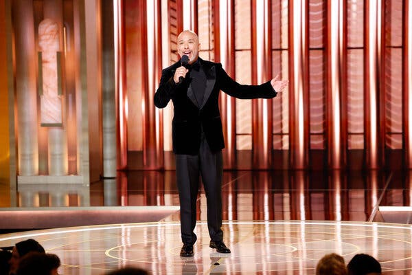Un hombre con esmoquin negro y camisa negra se encuentra en un escenario dorado brillante y habla por un micrófono.