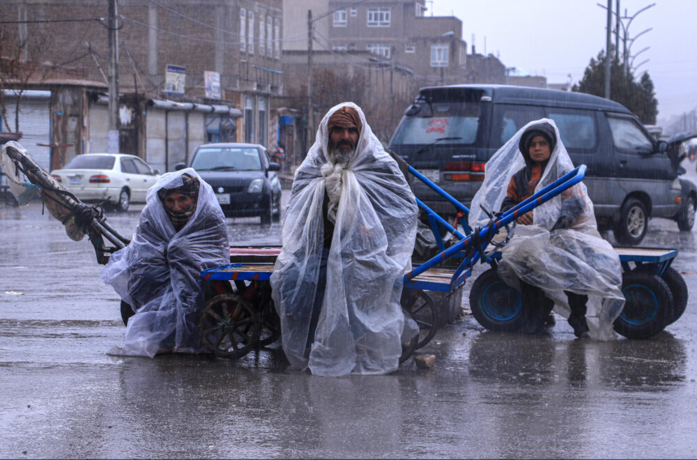 Unos trabajadores afganos se protegen de la lluvia con lonas de plástico mientras aguardan sentados sombre sus carritos en una calle de Herat, el 12 de marzo de 2024 al oeste de Afganistán