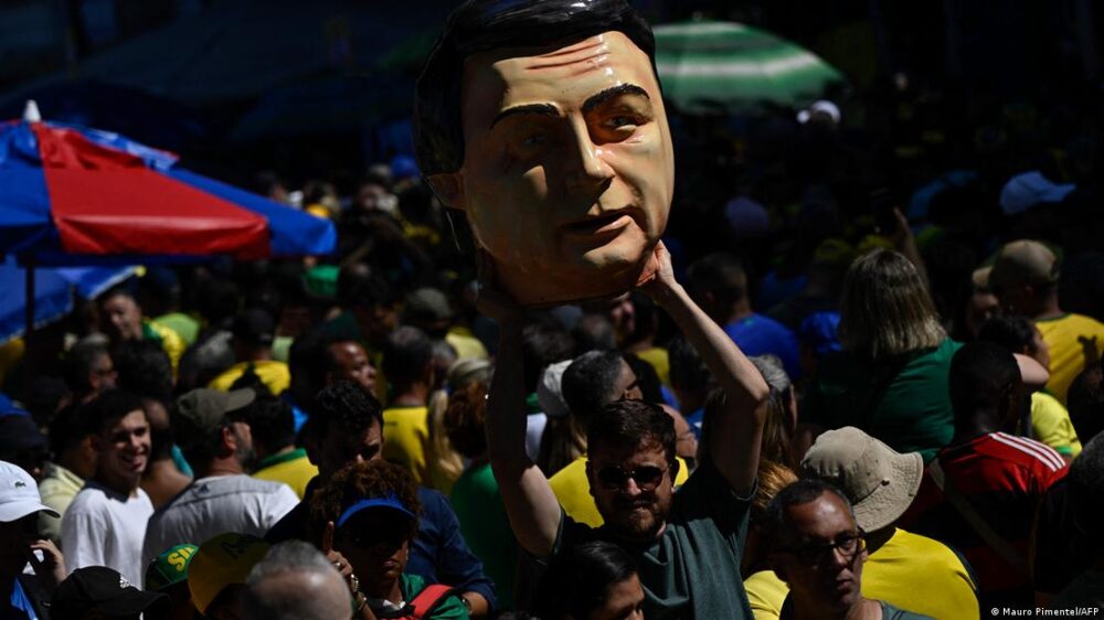 Un manifestante sostiene entre la multitud una grán máscara de Bolsonaro como de gigantes y cabezudos.