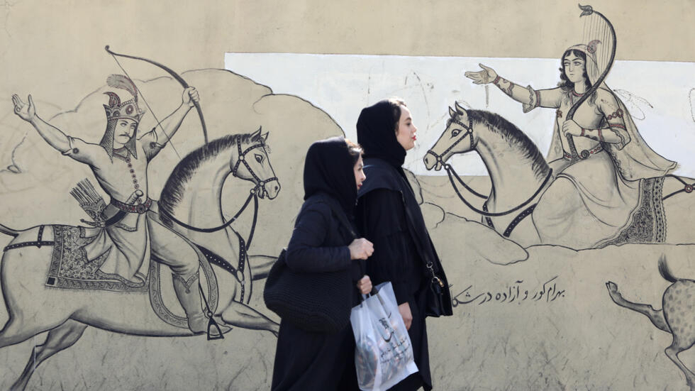 Dos mujeres con velo pasan por delante de un mural con una ilustración del poema épico persa "Shahnama" ("Libro de los reyes"), en el centro de Teherán, el 30 de enero de 2024