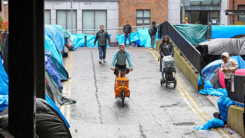 Unos peatones transitan junto al campamento de migrantes y solicitantes de asilo instalado en las cercanías de la Oficina de Protección Internacional, en Dublín, el 30 de abril de 2024