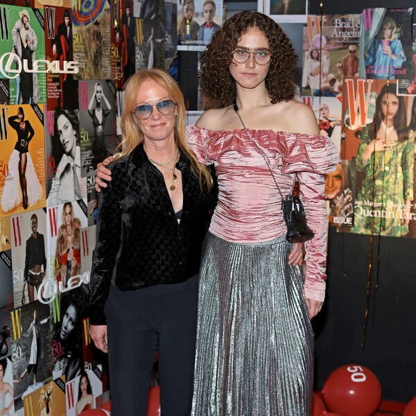 Kerstin Emhoff, wearing a black velvet shirt and black pants, stands next to Ella Emhoff, in an off-the shoulder long sleeve pink top and a pleated silver skirt. A backdrop of women’s magazine covers is behind them. 