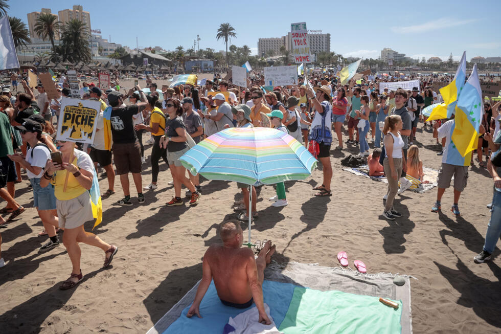 Una manifestación contra el turismo masivo pasa junto a un turista bajo una sombrilla en la playa canaria de Las Américas, el 20 de octubre de 2024 en Arona, en la isla española de Tenerife