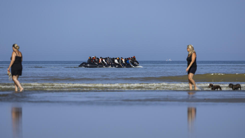 Dos turistas pasean por la orilla del mar mientras unos migrantes suben a la embarcación con la que intentarán cruzar el canal de la Mancha para llegar a suelo británico, el 30 de junio de 2025 en la playa de Neufchatel-Hardelot, al norte de Francia