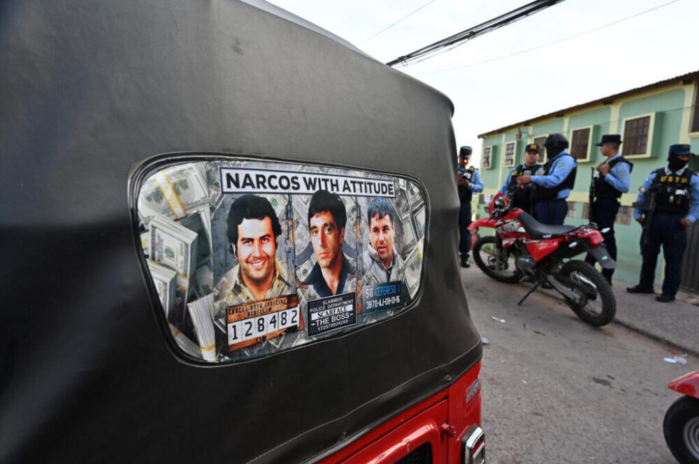 Retratos de los narcos Pablo Escobar y Joaquín "El Chapo" Guzmán, junto al del protagonista del filme "Caracortada" Tony Montana (Al Pacino), en una mototaxi mientras policías patrullan Tegucigalpa, el 14 de agosto de 2025, bajo estado de excepción.