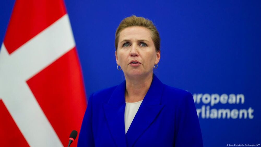 Primera ministra socialdemócrata danesa Mette Frederiksen, frente a un micrófono. De fondo, una bandera danesa y una pared azul del Parlamento Europeo.