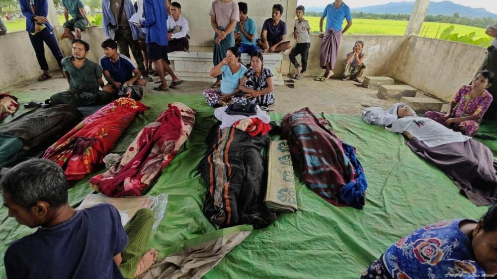 Varios cuerpos amortajados con telas yacen en el suelo mientras familiares y amigos permanecen a su alrededor.