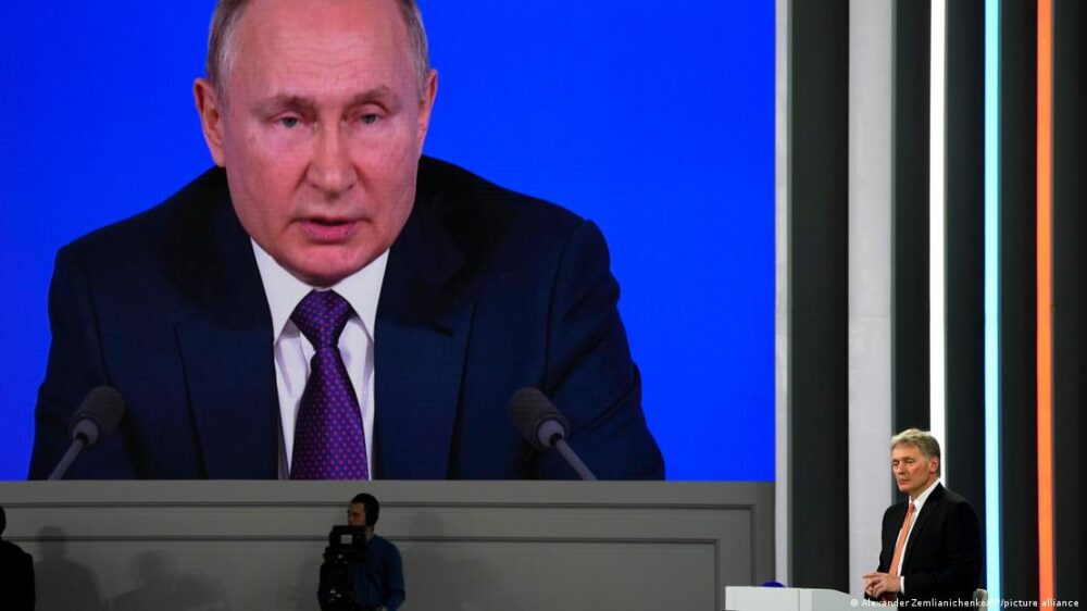 Vladimir Putin, presidente de Rusia, en una conferencia en Moscú, y el portavoz del Kremlin, Dmitri Peskov, en una tarima, escuchando a Putin.