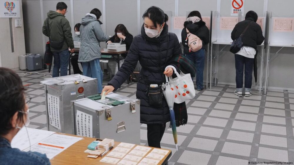 La mujer, con abrigo, tapabocas y paraguas, mete su voto en la urna. La mujer, con abrigo, tapabocas y paraguas, mete su voto en la urna.