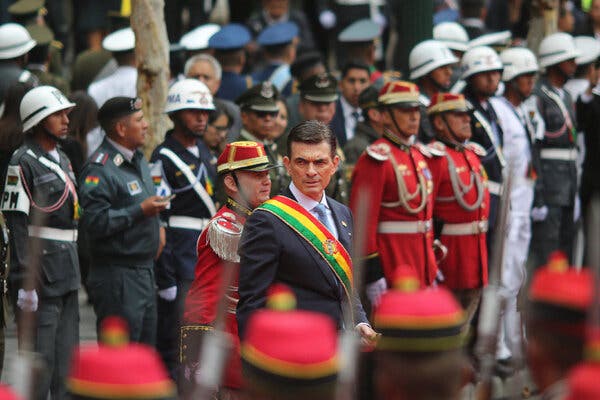 Un hombre vestido de traje y con una banda roja, amarilla y verde, de pie en medio de hombres con uniforme militar.