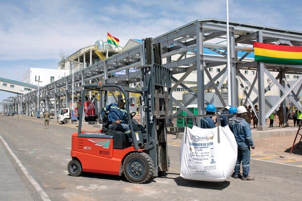 Una carretilla elevadora que transporta una gran bolsa blanca en una zona industrial, con unas cuantas personas con cascos de protección de pie alrededor.