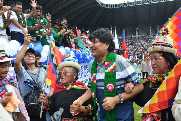 Un hombre vestido con una banda verde y flanqueado por dos mujeres que sostienen banderas de colores sonríe a un grupo de personas animadas, en un estadio.