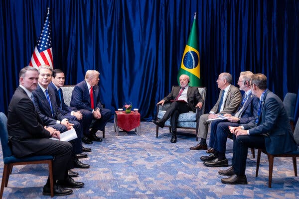 Personas trajeadas sentadas en círculo sobre una alfombra estampada, frente a frente. Dos personas con corbatas rojas están frente a frente en una mesa pequeña.