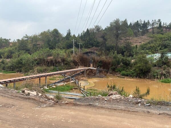 Un largo puente cruza un río naranja y fangoso. En primer plano hay un camino de tierra, con una colina cubierta de árboles y líneas eléctricas por encima.
