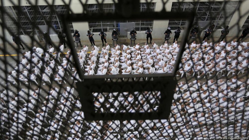 A través de una reja, presos sentados de forma perfectamente alineada. A través de una reja, presos sentados de forma perfectamente alineada.