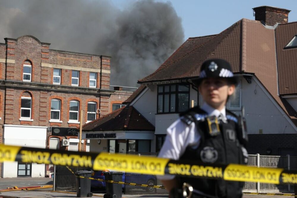 Una agente monta guardia en una zona acordonada por la policía tras el incendio en un supermercado del barrio de Golders Green, cuya humareda negra se eleva sobre unos edificios, el 27 de mayo de 2026 al norte de Londres